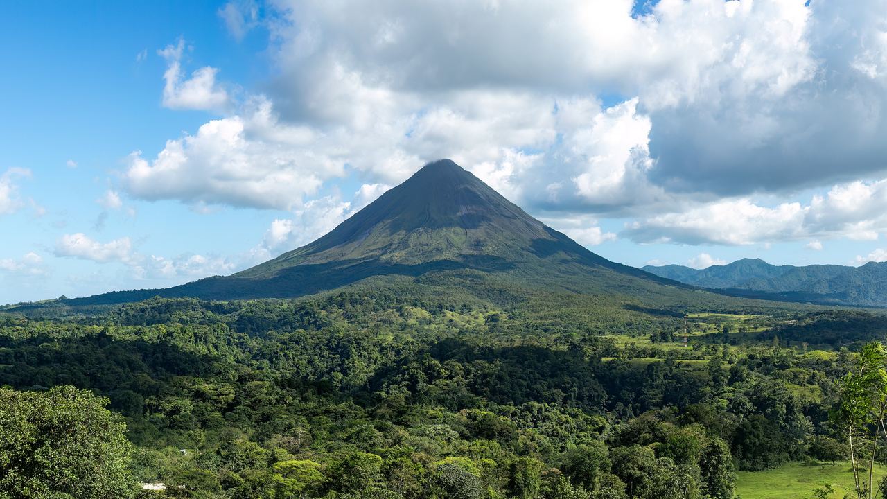 Wide view of the Arenal Volcano in Costa Rica, its green cone rising above rainforest with cloud gathering on the summit