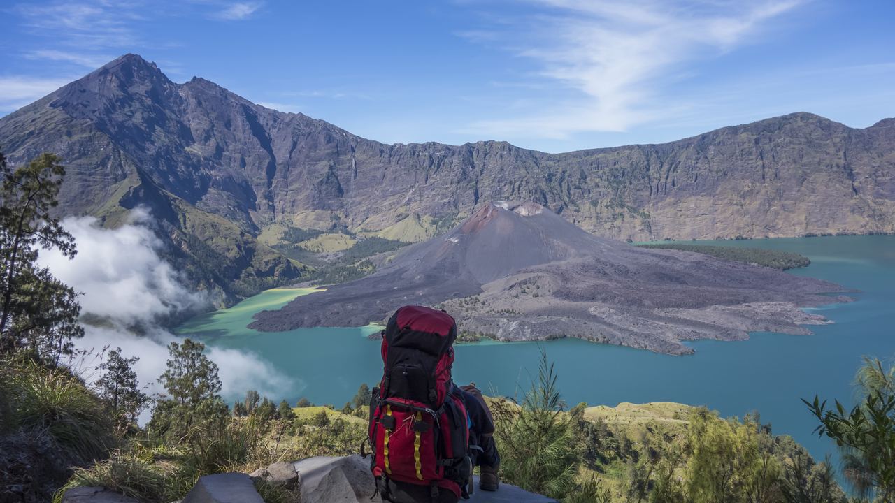 A trekker sitting on the crater rim of Mount Rinjani with a pack beside him, looking down at the Segara Anak lake and the Barujari cone