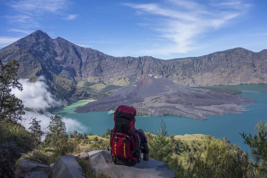 A trekker sitting on the crater rim of Mount Rinjani with a pack beside him, looking down at the Segara Anak lake and the Barujari cone