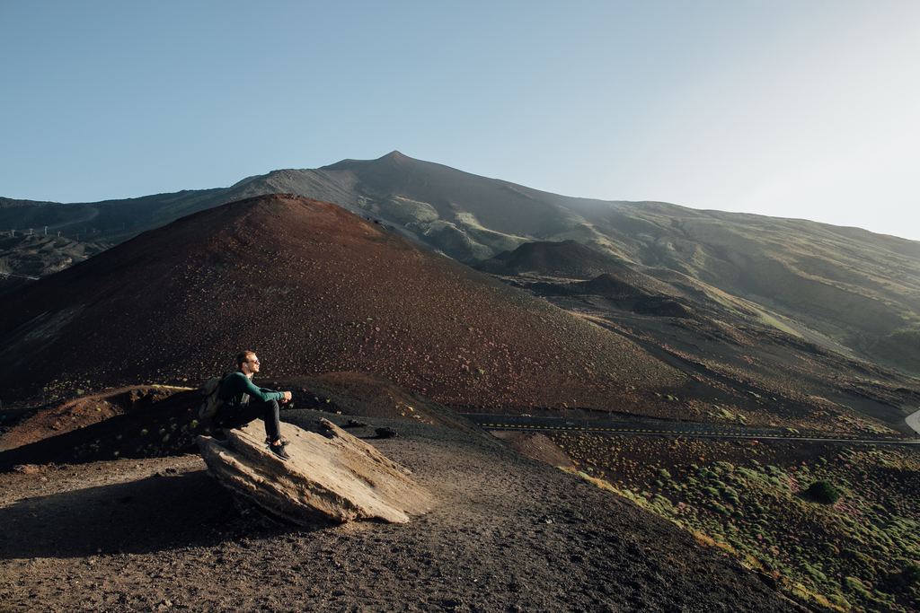 A walker sitting on lava rock high on Mount Etna, looking across the volcano's upper slopes