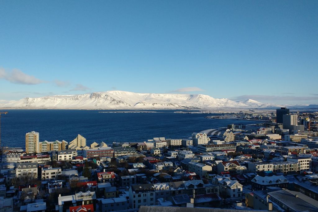 Aerial view of the coastal city of Reykjavík, Iceland, with snow-covered mountains behind it under a blue sky