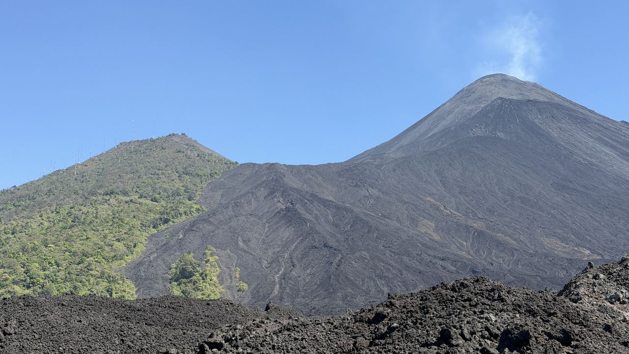The active MacKenney cone of the Pacaya degassing above a black lava field, with the wooded shoulder of the Cerro Grande on the left