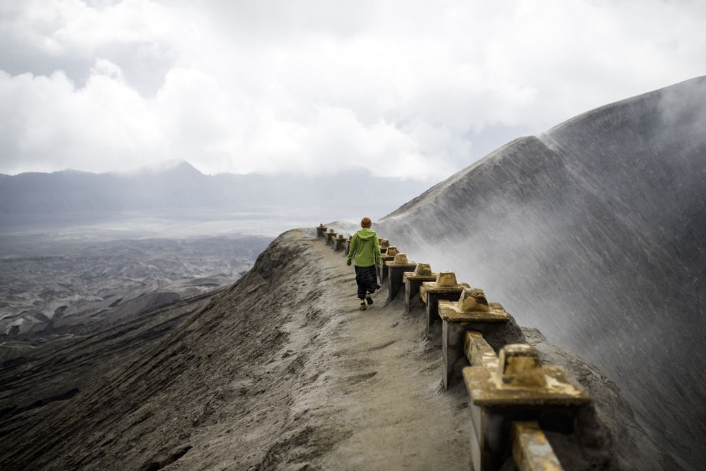 A visitor walking the narrow crater rim of Mount Bromo, with a handrail on one side and the open vent steaming on the other